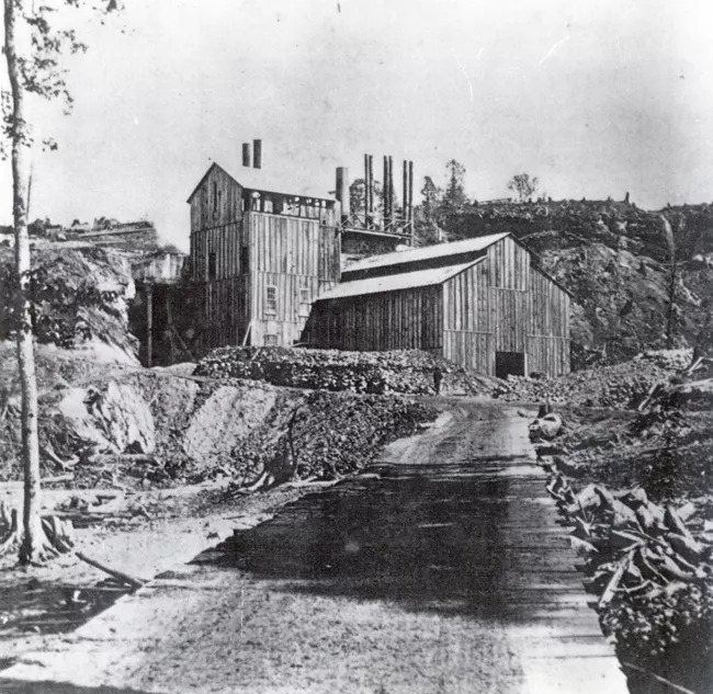 The Schoolcraft Blast Furnace at its original location on Munising creek. Munising Falls is visible in the distance at the upper right-hand corner. 